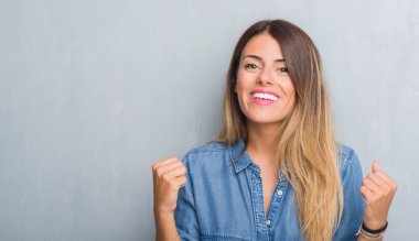Grunge gri duvar denim kıyafet kutluyor giyen genç yetişkin kadın şaşırttı ve başarı için kaldırdı kollarını açarak hayret ve gözleri açık. Kazanan kavramı.