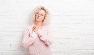 Sarışın genç kadın kıvırcık saçlı beyaz tuğla duvar ile gülümseyen üzerinde kapalı gözler ve yüzünde minnettar jest ile göğüs üzerinde eller. Sağlık kavramı.