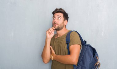 Üzerinde gri grunge duvar giyen genç adam yakışıklı turist sırt çantası çene düşünme hakkında soru, dalgın ifade üzerine el ile. Düşünceli yüzüyle gülümsüyor. Şüphe kavramı.
