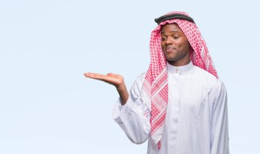 Geleneksel keffiyeh neşeli sunulması ve kameraya bakarak avucunun ile işaret gülümseyen izole arka plan üzerinde giyen genç Arapça Afrika adam.