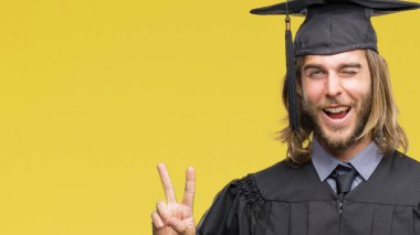 Genç yakışıklı adam uzun saçlı gülen yüz zafer işareti yaparken kameraya göz kırpıyor ile gülümseyen izole arka plan üzerinde mezun oldu. İki numaralı.