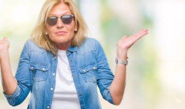 Orta yaş sarışın kadın izole arka plan clueless ve şaşkın bir ifadeyle kollar ve eller kaldırdı üzerinde güneş gözlüğü takıyor. Şüphe kavramı.