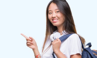 Sırt çantası ve kulaklık izole arka tarafına el ve parmak ile işaret çok mutlu üzerinde giyen genç Asyalı kadın