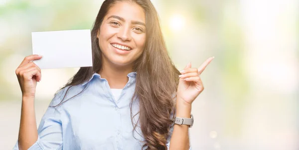 Young arab woman holding blank card over isolated background very happy pointing with hand and finger to the side