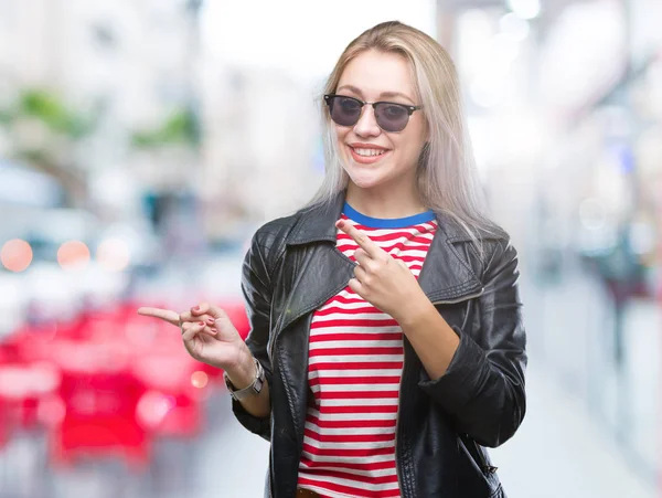 Young blonde woman wearing fashion jacket and sunglasses over isolated background smiling and looking at the camera pointing with two hands and fingers to the side.