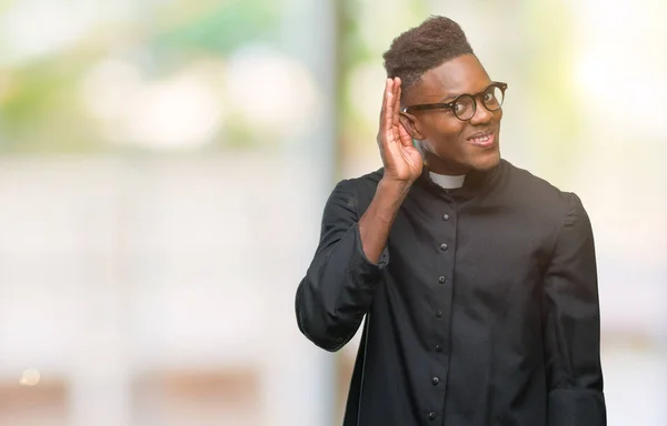 Young african american priest man over isolated background smiling with hand over ear listening an hearing to rumor or gossip. Deafness concept.
