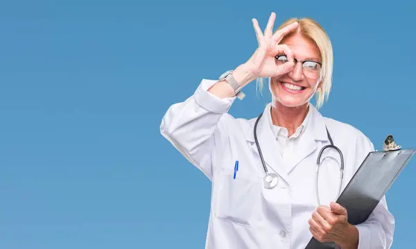 中年金发医生妇女持有剪贴板在孤立的背景与快乐的脸微笑做 标志与手在眼睛看通过手指 — 图库照片