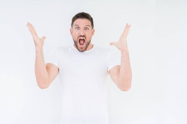 Üzerinde rahat beyaz t-shirt giyen genç yakışıklı adam deli kutluyor arka plan izole ve başarı için kaldırdı kollarını açarak hayrete ve heyecanlı çığlık gözleri açık. Kazanan kavramı