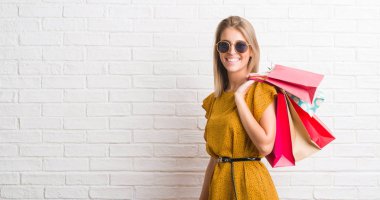 Güzel genç kadın ayakta ve diş gösterilen kendine güvenen bir gülümsemeyle gülümseyerek mutlu bir yüz ile satış alışveriş torbaları holding beyaz tuğla duvarın üzerinden