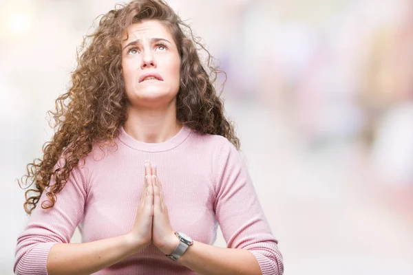 Beautiful brunette curly hair young girl wearing pink sweater over isolated background begging and praying with hands together with hope expression on face very emotional and worried. Asking for forgiveness. Religion concept.