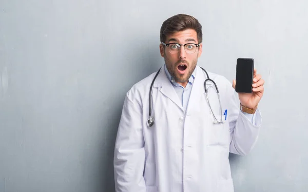 Handsome young doctor man over grey grunge wall holding smartphone scared in shock with a surprise face, afraid and excited with fear expression