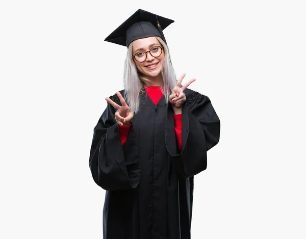 若いブロンドの女性は笑みを浮かべて勝利のサインをやって指を表示するカメラを分離の背景に大学院の制服を着ています — ストック写真
