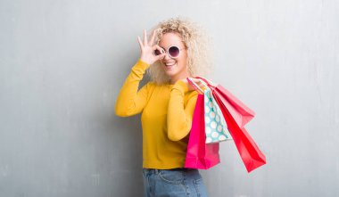 Genç Sarışın kadın grunge yüzünden mutlu yüz el parmaklarının arasından seyir göz üzerinde ok işaretiyle yapıyor gülümseyen ile satış alışveriş torbaları holding arka plan gri