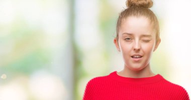 Bun ve seksi bir ifadeyle, neşeli ve mutlu yüzünü kameraya bakarak göz kırpıyor kırmızı kazak giyen genç sarışın kadın.