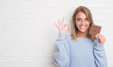 Güzel ve genç birbayan yeme çikolata bar ok işareti parmaklar, mükemmel sembolü ile yapıyor beyaz tuğla duvarın üzerinden
