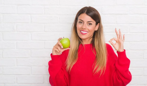 年轻的成年妇女在白色砖墙吃新鲜的绿色苹果做确定的标志与手指 优秀的象征 — 图库照片