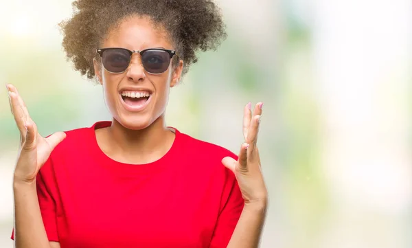Young afro american woman wearing sunglasses over isolated background crazy and mad shouting and yelling with aggressive expression and arms raised. Frustration concept.