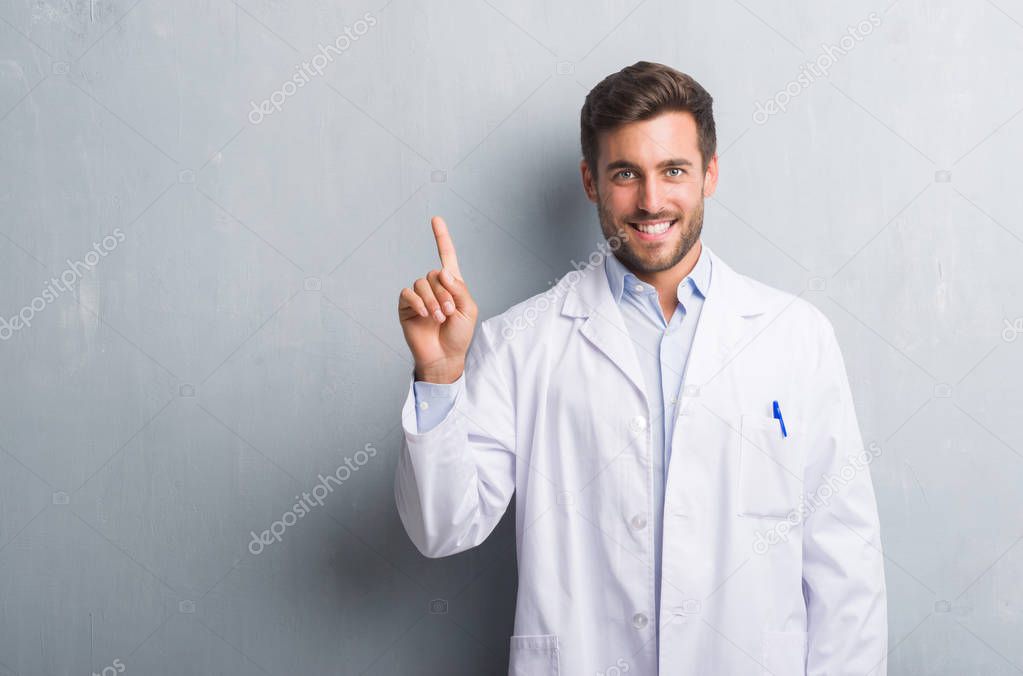 Handsome young professional man over grey grunge wall wearing white coat showing and pointing up with finger number one while smiling confident and happy.