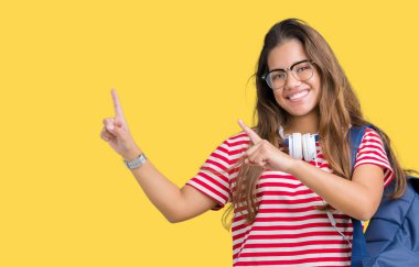 Gülümseyen ve iki elleri ve parmakları ile tarafına işaret kamera bakarak izole arka plan üzerinde kulaklık ve sırt çantası giyen genç güzel esmer öğrenci kadın.