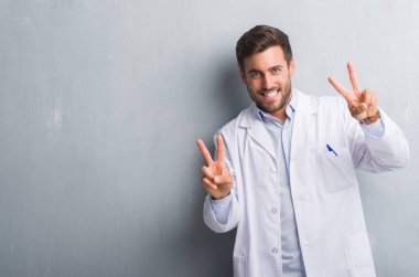 Yakışıklı genç profesyonel adam beyaz giyen gri grunge duvarın üzerinden kat gülümseyen zafer işareti yapıyor parmak gösterilen kamera arıyorum. İki numaralı.