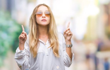 Genç güzel sarışın kadın hayrete ve yukarıya bakıyor ve parmakları ile işaret ve silah kaldırdı sürpriz izole arka plan üzerinde güneş gözlüğü takıyor.