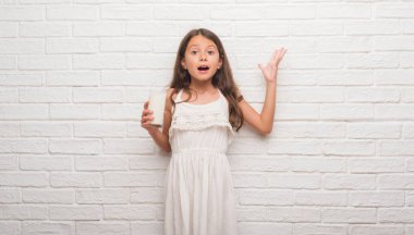 Genç İspanyol bir bardak süt çok mutlu ve heyecanlı, kazanan ifade büyük gülümseme ile çığlık zaferini kutluyor içme Beyaz tuğla duvarın üzerinden çocuk ve ellerini kaldırdı