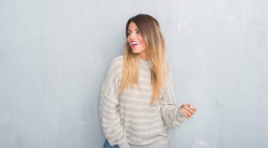 Grunge gri duvar uzak tarafına yüzünde, doğal ifade gülümseme ile seyir kış kazak giyen genç yetişkin kadın. Kendine güvenen gülüyor.