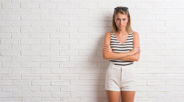 Genç güzel sarışın kadın beyaz tuğla duvar şüpheci ve çapraz kolları olan yüzündeki gergin, onaylamayan ifade yüzünden. Negatif kişi.