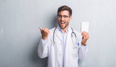 Yakışıklı genç doktor adam işaret ve başparmak kadar mutlu yüz gülümseyen tarafı ile gösterilen boş defter tutan gri grunge duvarın üzerinden