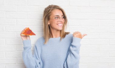Güzel genç kadın işaret ve başparmak kadar mutlu yüz gülümseyen tarafı ile gösterilen pizza dilimi yemek beyaz tuğla duvarın üzerinden