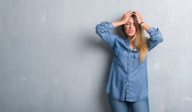 Genç Yetişkin kadın çünkü umutsuz ve stresli baş ağrısı acı denim kıyafeti giymiş grunge gri duvar yüzünden acı ve migren. Ellerini kafasına.