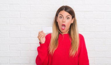 Genç Yetişkin kadın boş tutan beyaz tuğla duvarın üzerinden ziyaret kart bir sürpriz yüz, korkuyor ve heyecanlı korku ifadesi ile şok korkmuş