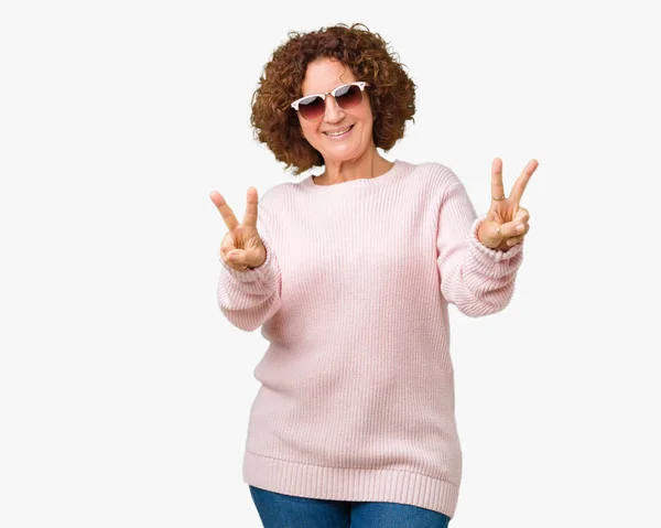 美丽的中年老年妇女穿着粉红色的毛衣和太阳镜在孤立的背景微笑着看着相机显示手指做胜利的标志 — 图库照片