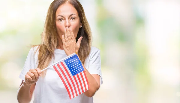 Middle age hispanic woman holding flag of United States of America over isolated background cover mouth with hand shocked with shame for mistake, expression of fear, scared in silence, secret concept