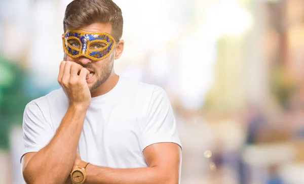Young handsome man wearing carnival mask over isolated background looking stressed and nervous with hands on mouth biting nails. Anxiety problem.
