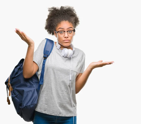 若いアフロ アメリカ学生女性を身に着けているヘッドフォンとバックパックは 腕と手を上げて背景無知と混乱式を分離しました 疑いのコンセプト — ストック写真