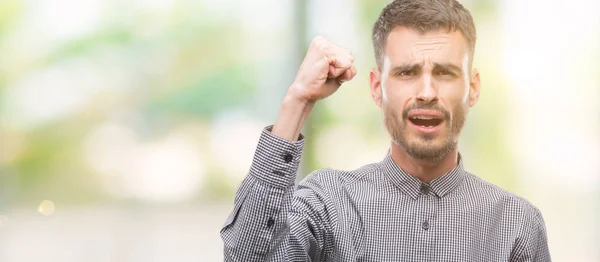 Young hipster man angry and mad raising fist frustrated and furious while shouting with anger. Rage and aggressive concept.