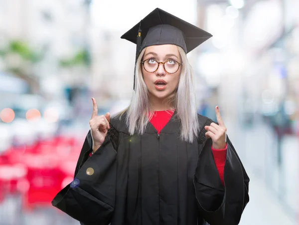 若いブロンドの女性隔離された背景に大学院の制服を着てびっくりし 驚き見て 指で指して 腕を上げた — ストック写真