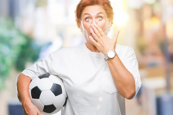 Atrractive senior caucasian redhead woman holding soccer ball over isolated background cover mouth with hand shocked with shame for mistake, expression of fear, scared in silence, secret concept