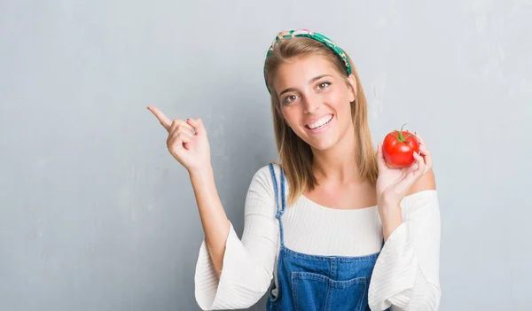 美丽的年轻女子在咕黑灰色的墙吃新鲜的西红柿非常高兴的指向手和手指的一侧 — 图库照片