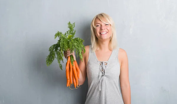 白种人成年妇女在灰色的沙哑墙上拿着新鲜的胡萝卜 一张幸福的脸站着 带着自信的微笑露出了牙齿 — 图库照片