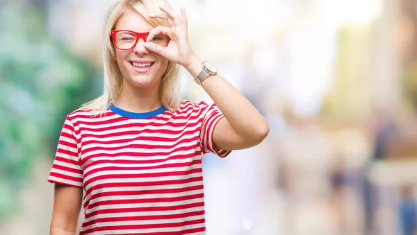 Young beautiful blonde woman wearing glasses over isolated background doing ok gesture with hand smiling, eye looking through fingers with happy face.