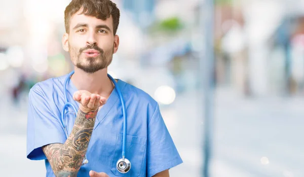 Young handsome nurse man wearing surgeon uniform over isolated background looking at the camera blowing a kiss with hand on air being lovely and sexy. Love expression.