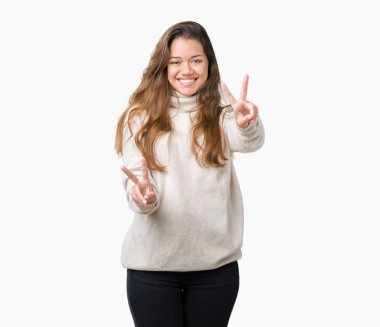 Zafer işareti yapıyor parmak gösterilen kamera seyir gülümseyen izole arka plan üzerinde boğazlı kazak giyen genç güzel esmer kadın. İki numaralı.