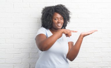 Genç Afro-Amerikan artı boyutu kadın hayrete ve el ile sunulması ve parmak ile işaret ederken kameraya gülümseyen beyaz tuğla duvarın üzerinden.