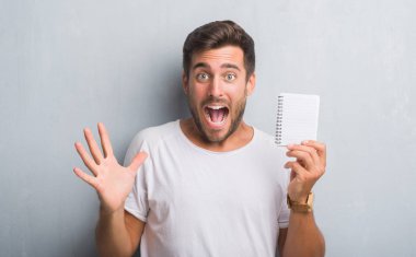 Yakışıklı genç gri grunge üzerinde duvar büyük gülümseme ile çığlık zaferini kutluyor ve ellerini kaldırdı boş defter çok mutlu ve heyecanlı, kazanan ifade holding
