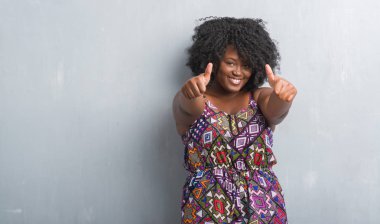 Gri grunge duvar onaylama renkli elbise giyen genç Afro-Amerikan kadın el ile pozitif jest yapıyor gülümseyen ve başarı için mutlu yaşasın. Kamera, kazanan jest arıyorsunuz.