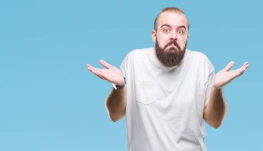 Üzerinde rahat t-shirt giyen genç beyaz hipster adam arka plan clueless ve şaşkın bir ifadeyle kollar ve eller kaldırdı izole. Şüphe kavramı.