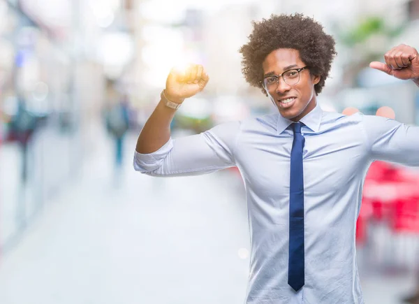 Afro american business man wearing glasses over isolated background showing arms muscles smiling proud. Fitness concept.