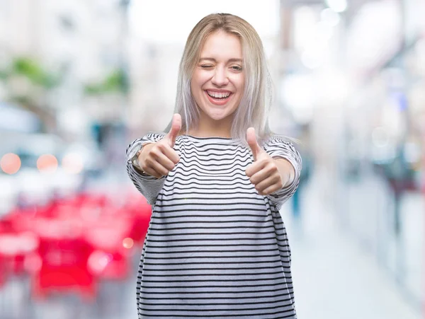 Young blonde woman over isolated background approving doing positive gesture with hand, thumbs up smiling and happy for success. Looking at the camera, winner gesture.